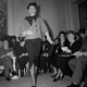 A woman walks in a black and white checkered, flamed jacket with matching gaiters. Under the jacket is a black wool dress, its narrow skirt shorter than those in many collections of the time and split in front. A black satin bow and a black beret complete the ensemble.